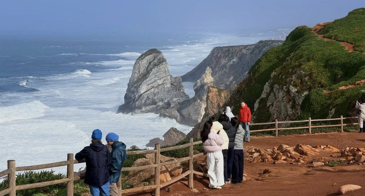 Visitors at a wooden fence watching surf break below green cliffs